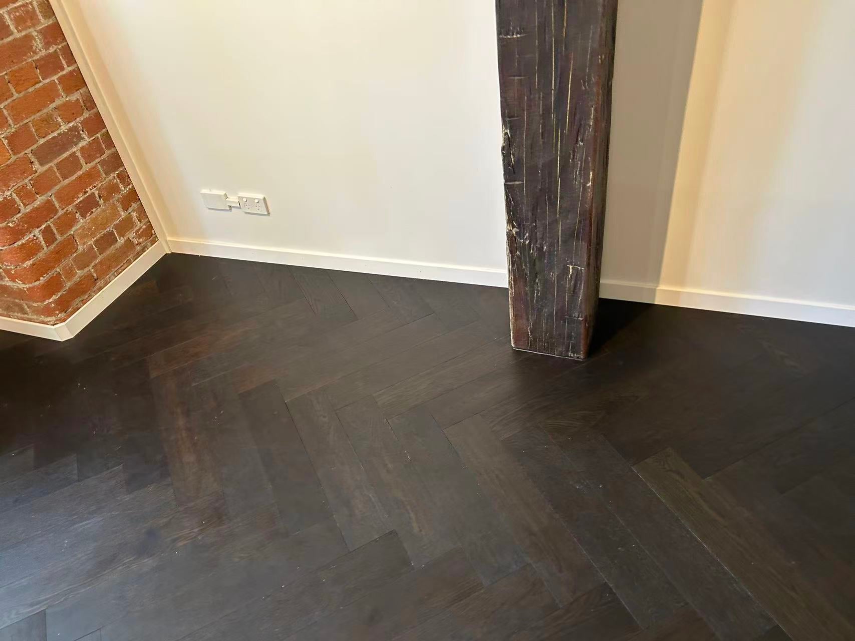 Midnight Grey iDesign Herringbone Flooring 15/3mm installed in a loft-style room featuring dark chevron timber planks, exposed red brick wall, and a rustic timber post.