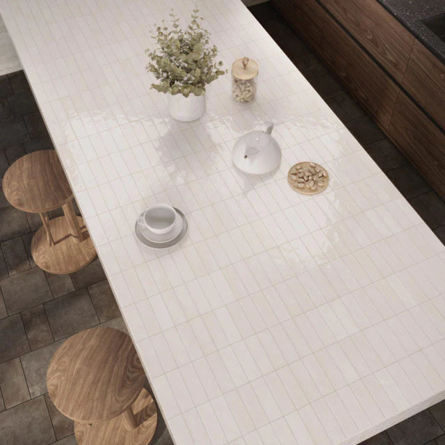 Overhead view of Mini Pure White Glossy 50x150mm tiles forming a sleek kitchen island countertop styled with wooden stools, a white teapot, and a potted plant.