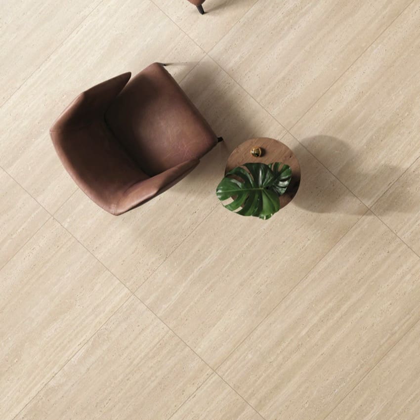 Tallow Vein Cut Travertine Look Beige 600x600mm porcelain floor tiles installed in a modern living room with a brown leather chair and round wooden side table with monstera plant.