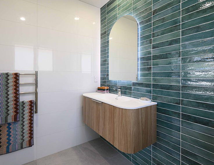 Mojave Sea Water Brick 60x250mm glossy teal vertical stacked tiles on a bathroom feature wall behind a floating timber vanity with arch backlit mirror.