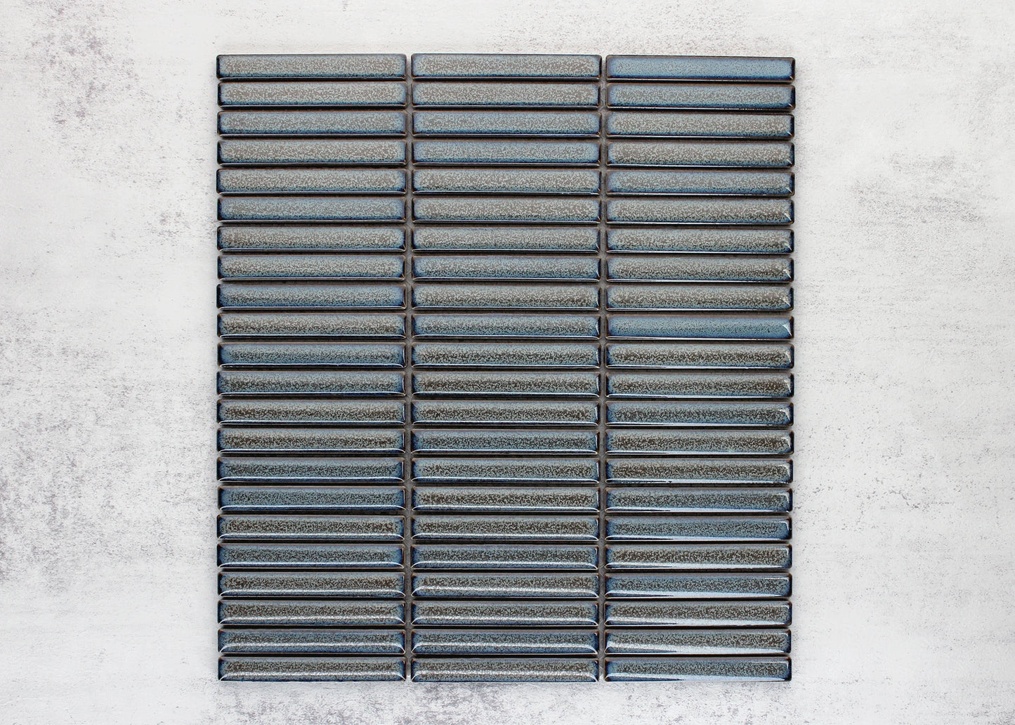 Flat lay pack shot of Mottled Grey Gloss Porcelain Glazed KitKat 12x92mm tiles arranged in three stacked columns showing blue-grey reactive glaze finish.