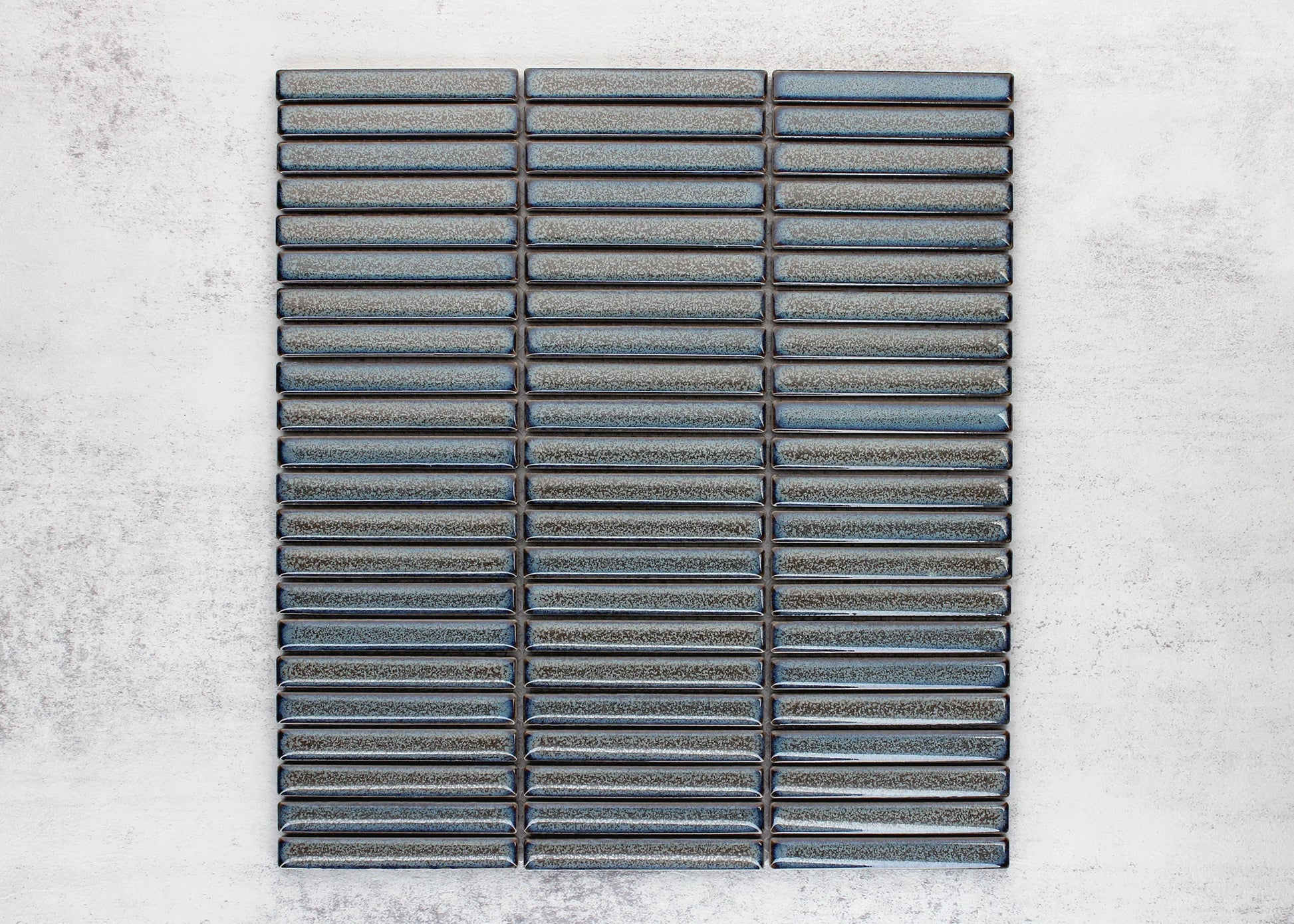 Flat lay pack shot of Mottled Grey Gloss Porcelain Glazed KitKat 12x92mm tiles arranged in three stacked columns showing blue-grey reactive glaze finish.