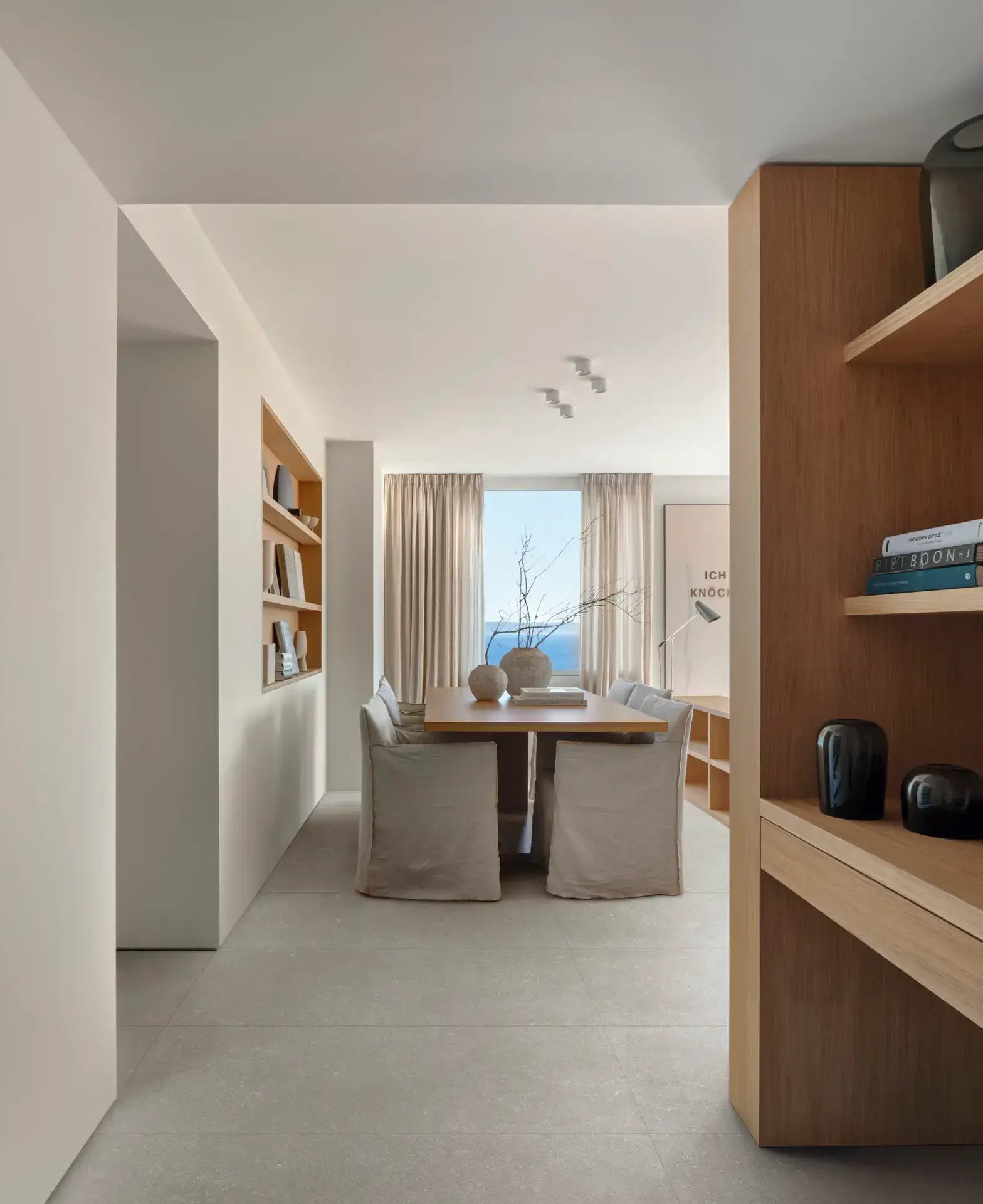 Mystone Berici Grigio Naturale 600x1200mm tiles laid on a dining room floor with linen chairs, wooden shelving, and a large ocean-view window.