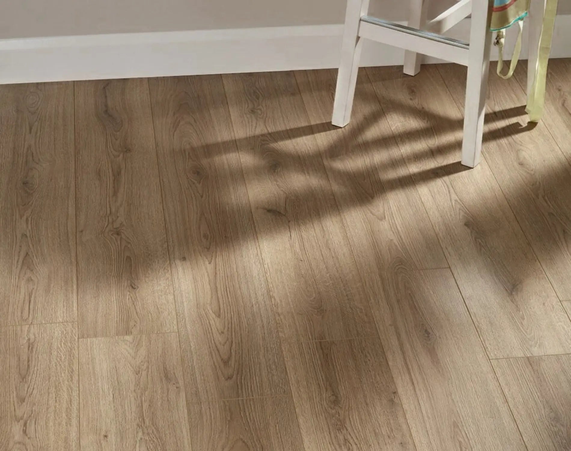 Natural Oak Urban Laminate 12mm Flooring 12mm installed in a bright room with warm sunlight casting shadows from a white chair across the wide-plank wood-effect surface.