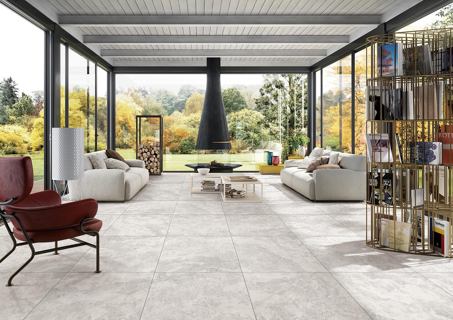 New Travertino Ivory Matt Rect 600x600mm tiles covering a bright modern living room floor with modular sofas, a suspended fireplace, and floor-to-ceiling garden views.