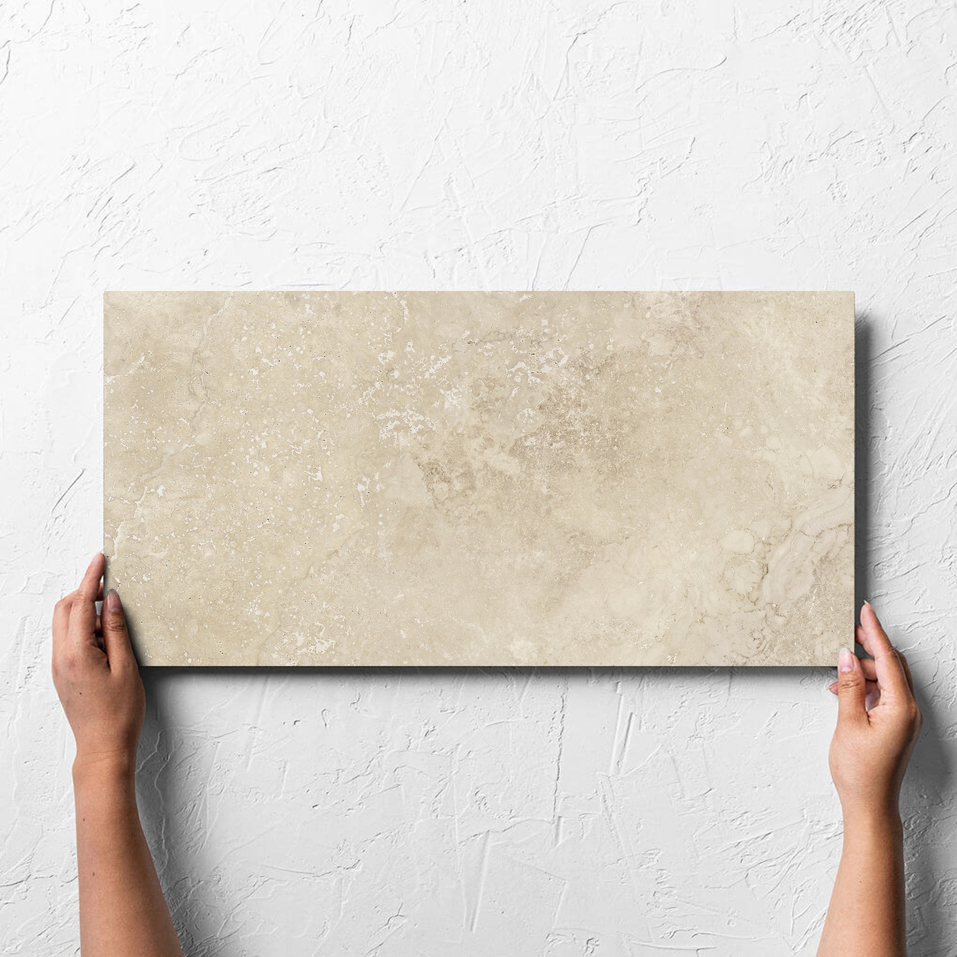 Hand holding a single Norcia Travertine Beige 300x600mm tile against a white rendered wall, showing the beige matt surface