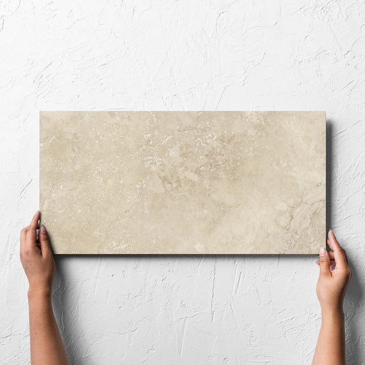 Hand holding a single Norcia Travertine Beige 300x600mm tile against a white rendered wall, showing the beige matt surface