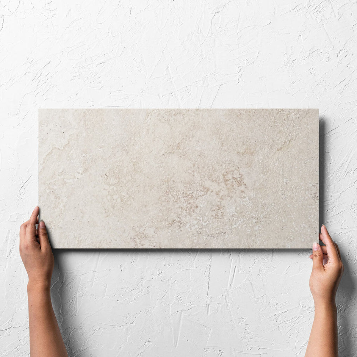 Hand holding a single Norcia Travertine Beige 300x600mm tile against a white rendered wall, showing the beige travertine surface