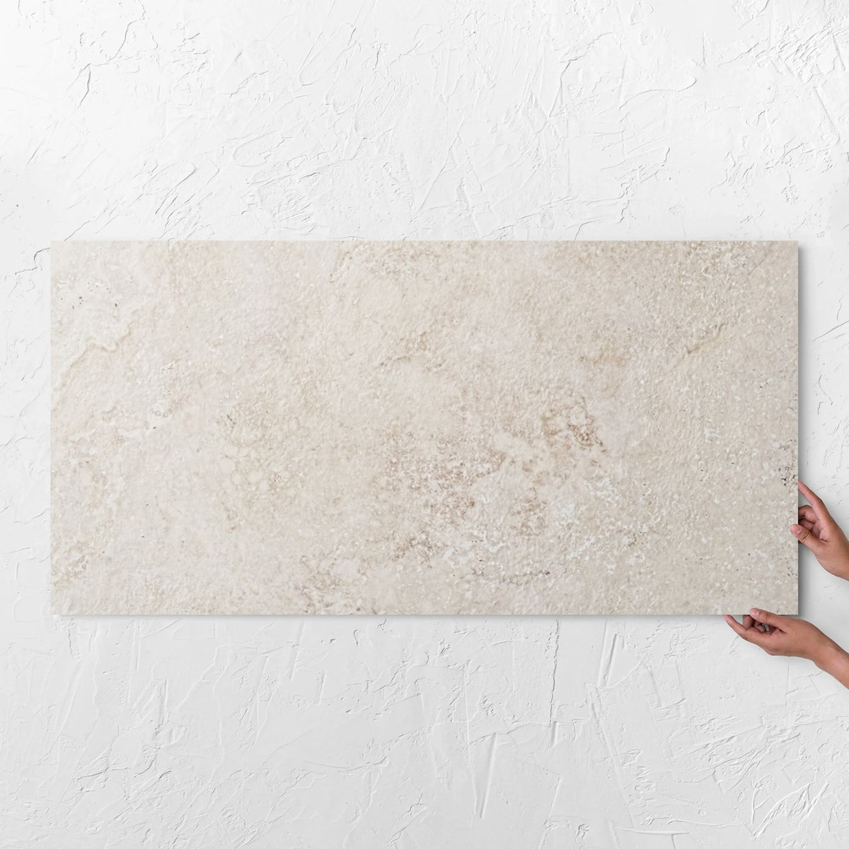 Hands holding a Norcia Travertine Beige 600x1200mm tile against a white rendered wall, showing the beige travertine surface