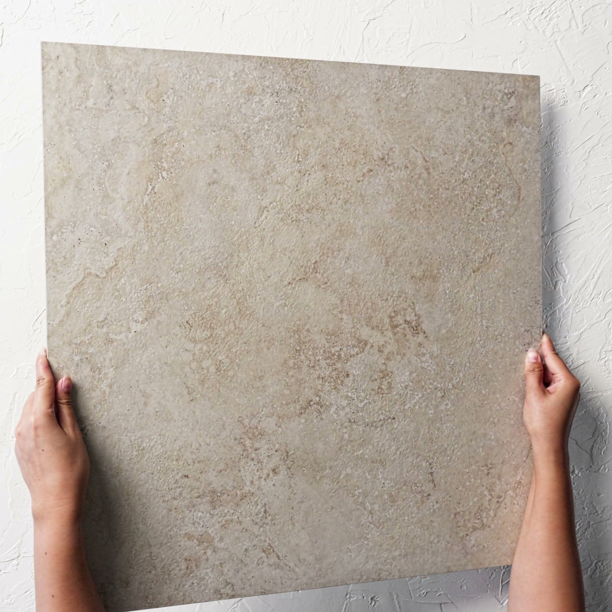 Norcia Travertine Beige External Rectified Paver 600x600x20mm held at an angle against a white rendered wall, showing the beige travertine surface