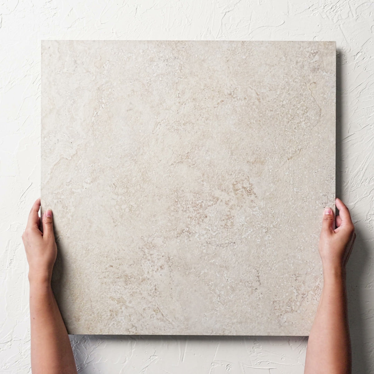 Hands holding a single Norcia Travertine Beige External Rectified Paver 600x600x20mm against a white rendered wall, showing the beige travertine surface