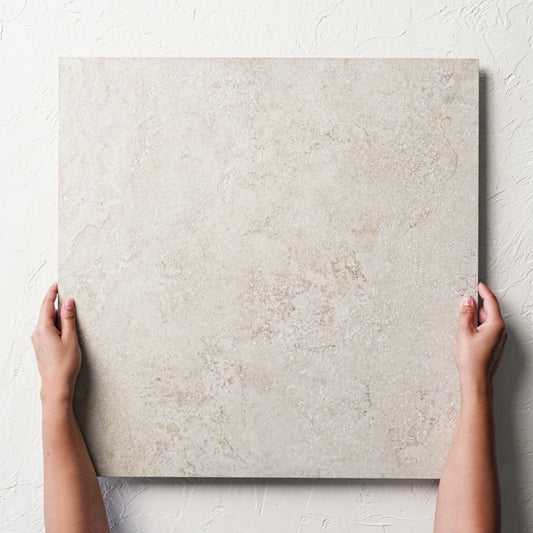Hands holding a single Norcia Travertine Beige External Rectified Paver 600x600x20mm against a white rendered wall, showing the beige travertine surface