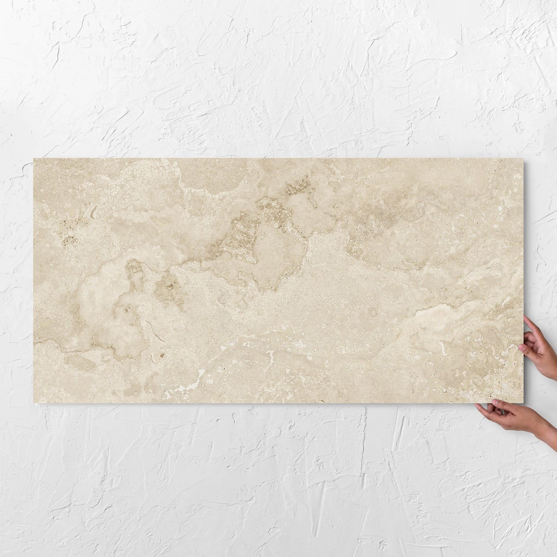 Hands holding a Norcia Travertine Beige Honed 600x1200mm tile against a white rendered wall, showing the tile surface
