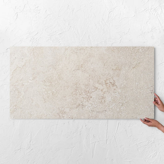 Hands holding a Norcia Travertine Beige Honed 600x1200mm tile against a white rendered wall, showing the beige travertine surface