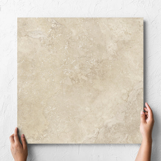 Hand holding a single Norcia Travertine Beige Honed 600x600mm tile against a white rendered wall, showing the tile surface