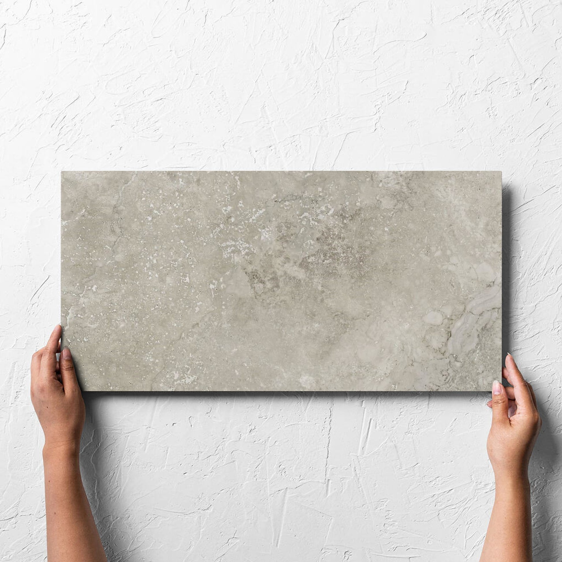 Hand holding a single Norcia Travertine Grey 300x600mm tile against a white rendered wall, showing the tile surface