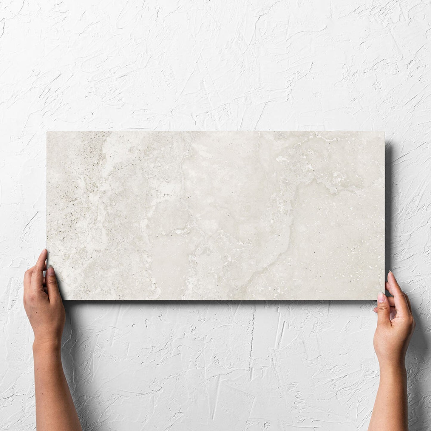 Hand holding a single Norcia Travertine Light 300x600mm tile against a white rendered wall, showing the tile surface