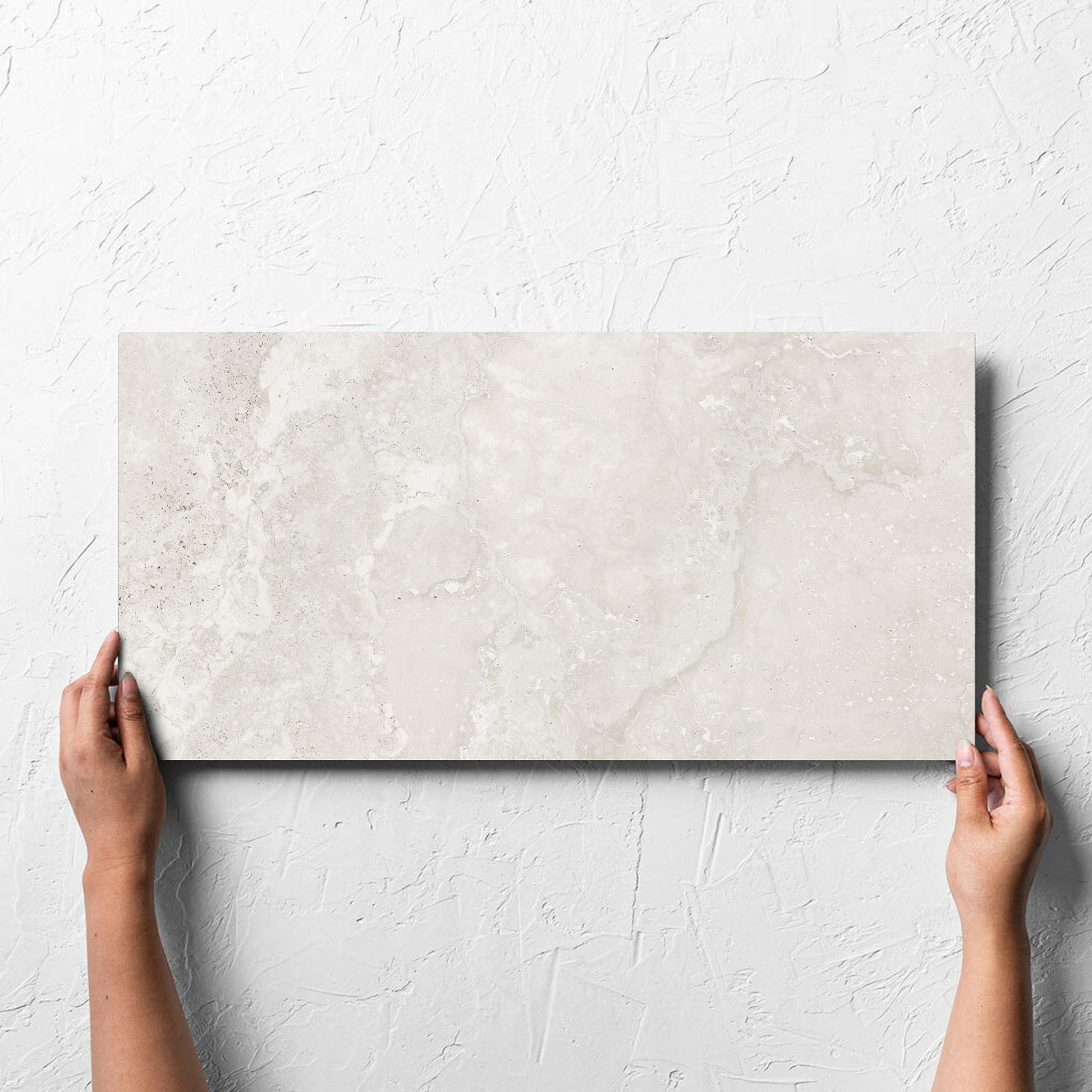 Hand holding a single Norcia Travertine Light 300x600mm tile against a white rendered wall, showing the tile surface