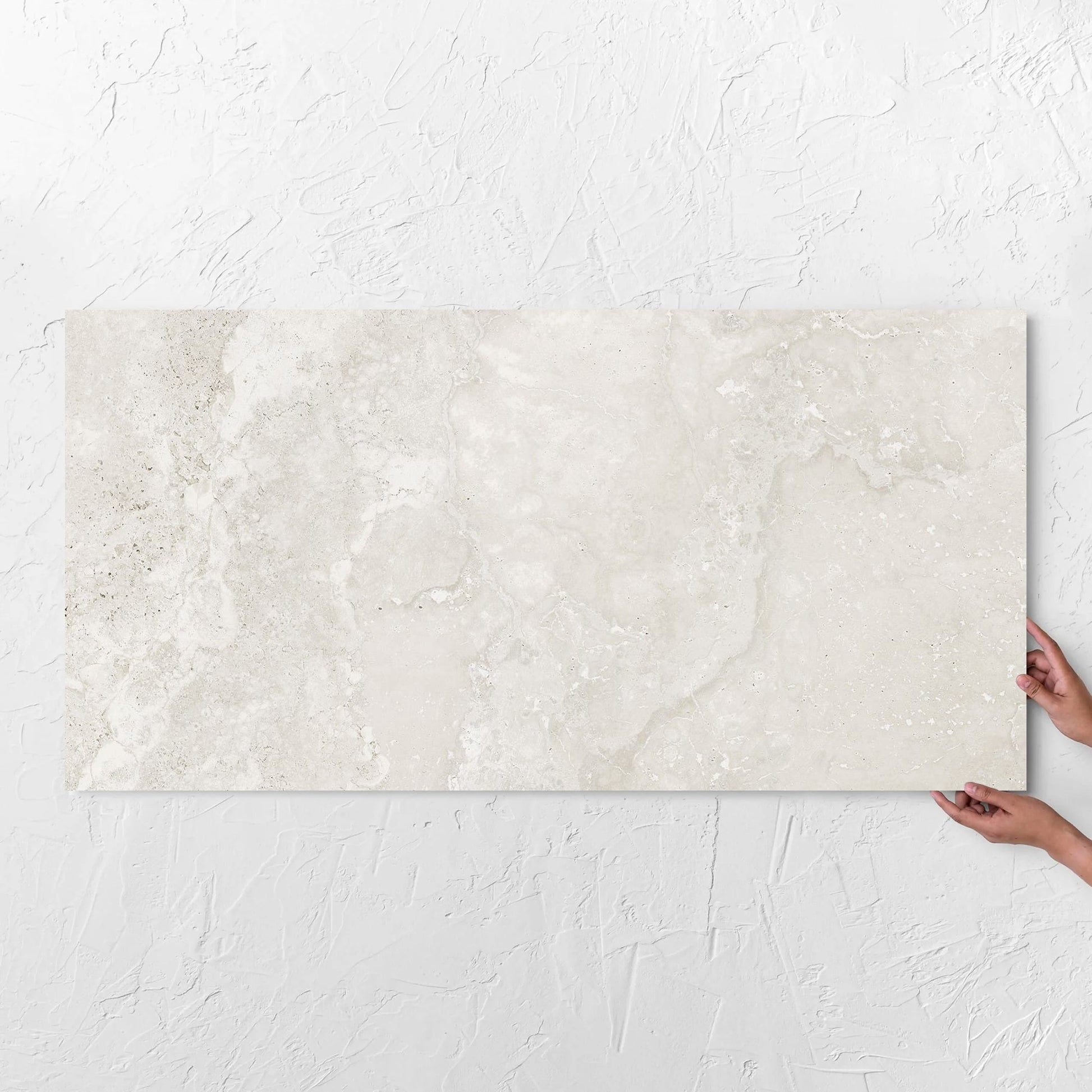 Hands holding a Norcia Travertine Light Bamboo Feature 600x1200mm tile against a white rendered wall, showing the tile surface
