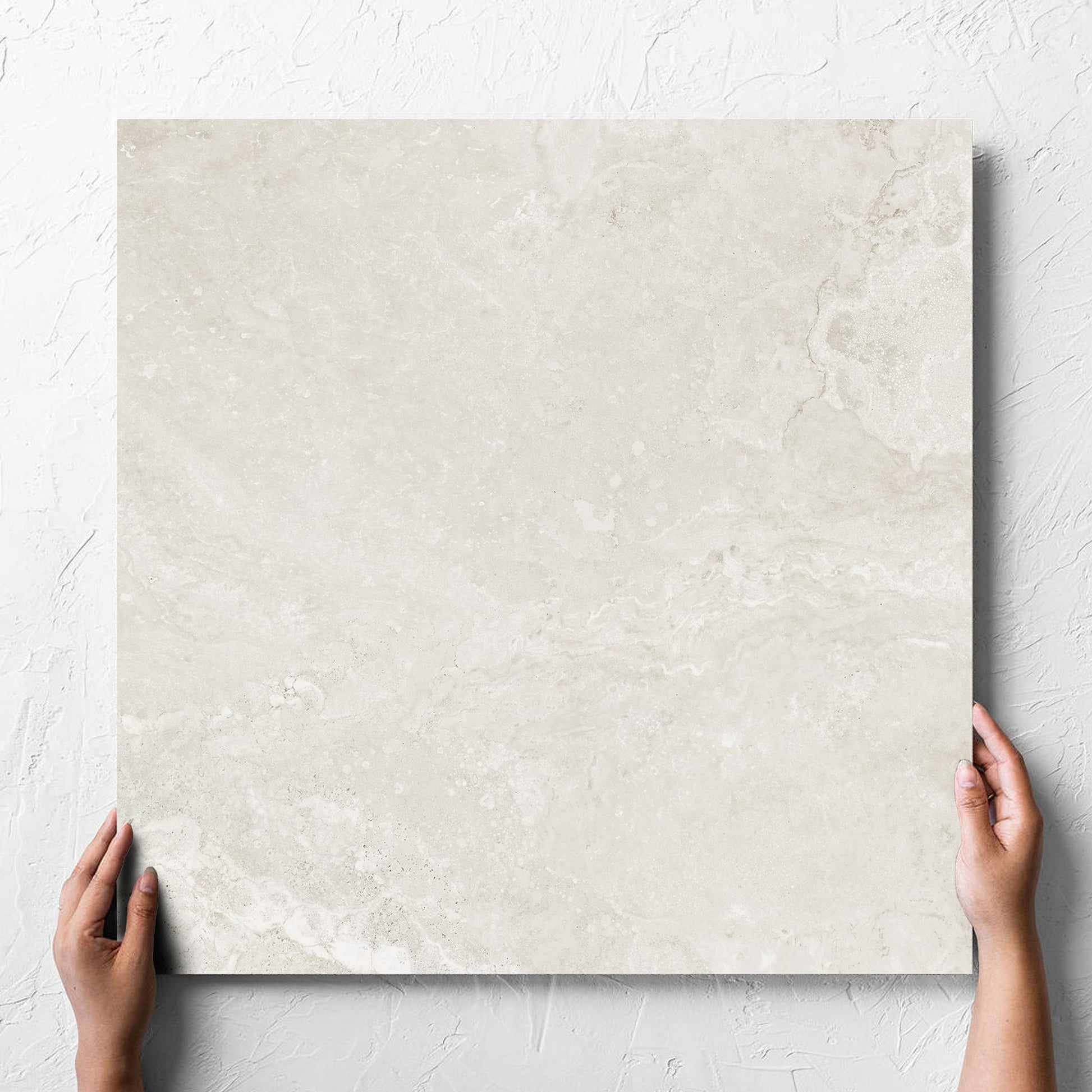 Hand holding a single Norcia Travertine Light Honed 600x600mm tile against a white rendered wall, showing the tile surface