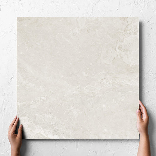 Hand holding a single Norcia Travertine Light Honed 600x600mm tile against a white rendered wall, showing the tile surface