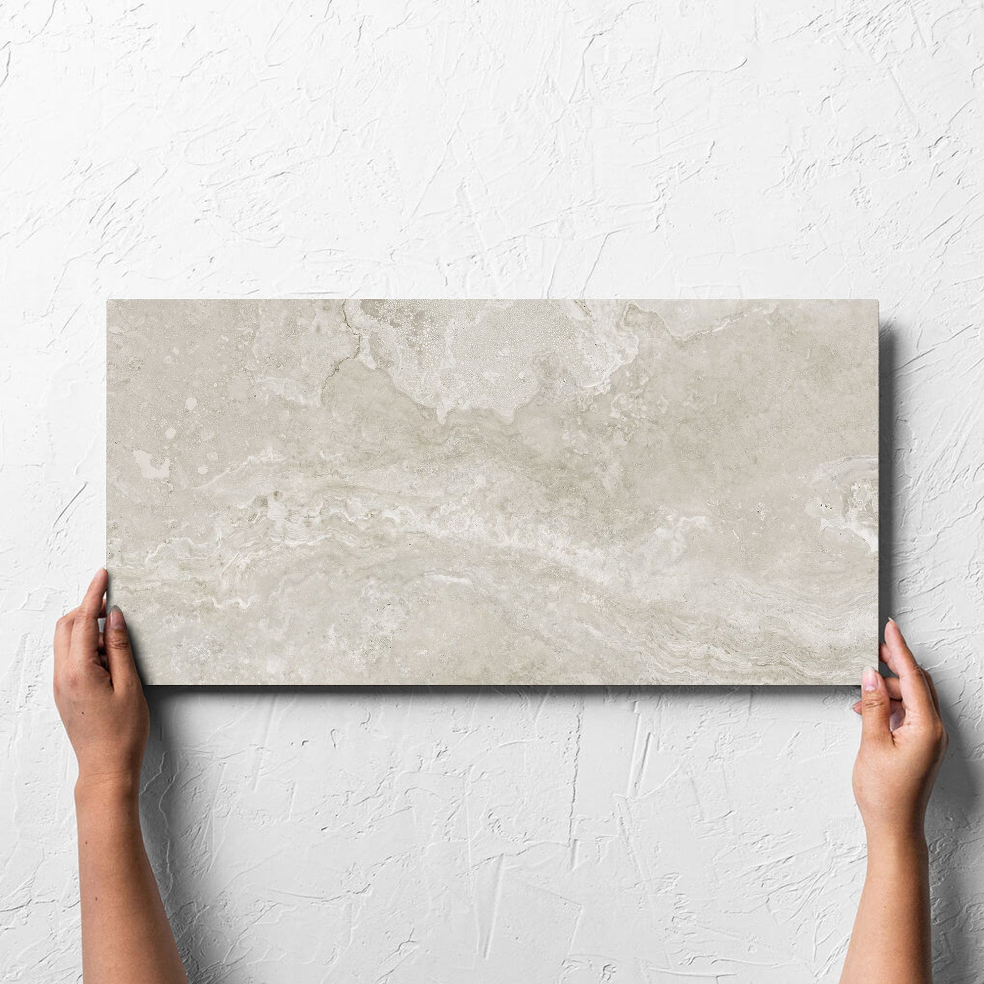 Hand holding a single Norcia Travertine Silver 300x600mm tile against a white rendered wall, showing the tile surface