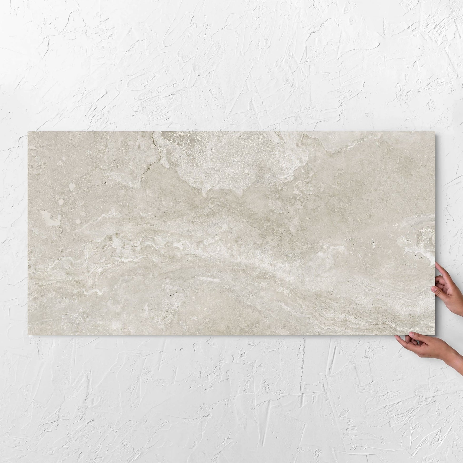 Hands holding a Norcia Travertine Silver 600x1200mm tile against a white rendered wall, showing the tile surface