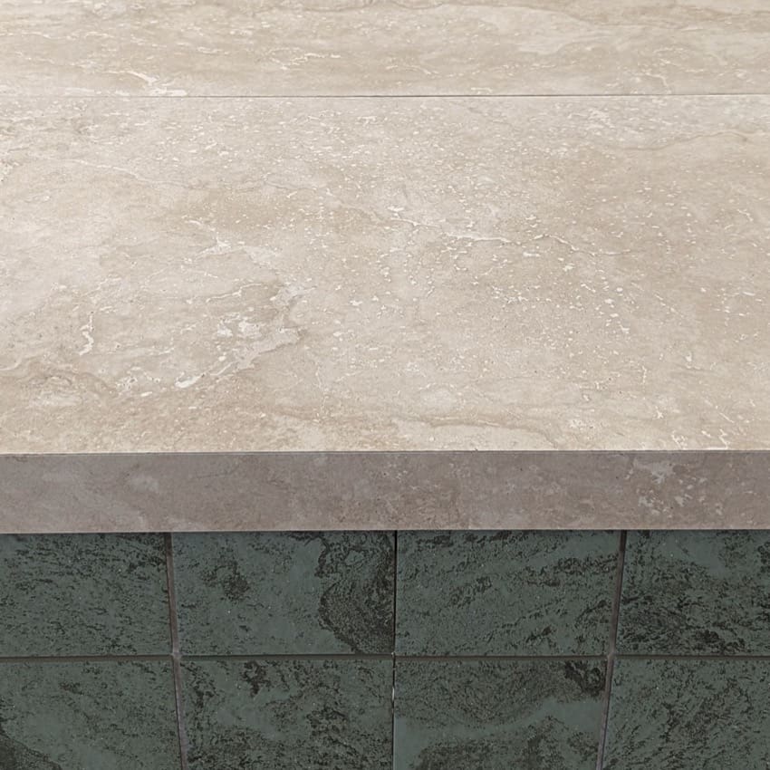 Norcia Travertine Silver 75x300mm tile used as a coping stone ledge over dark green ceramic wall tiles, showing natural beige veining and smooth matte finish.