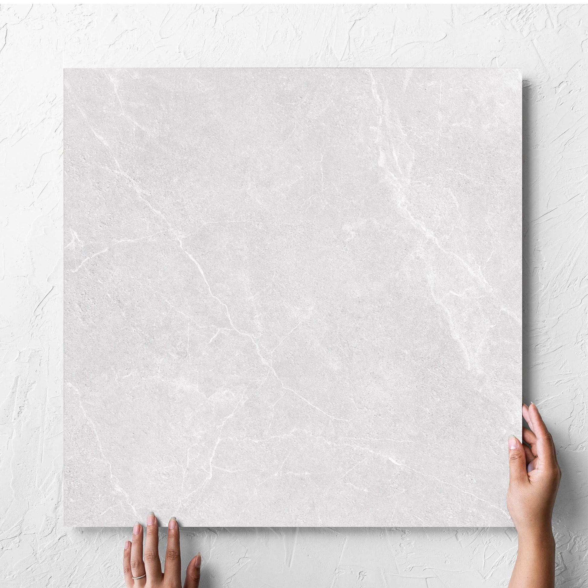 Hand holding a single Oglio Silver 600x600mm tile against a white rendered wall, showing the silver surface