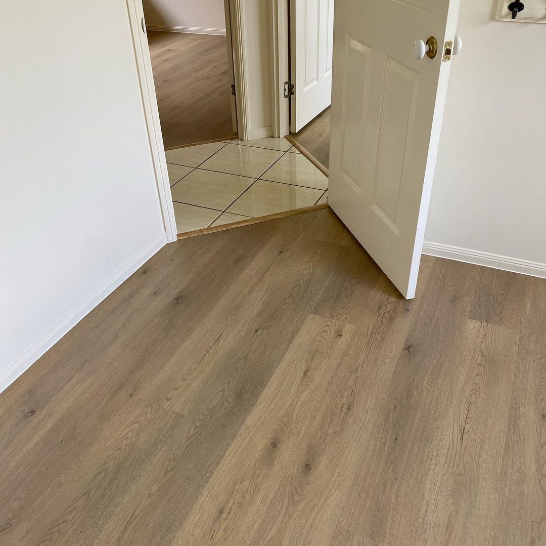 Pale Sand Elegant SPC Hybrid 6.5mm Flooring 5+1.5mm installed in a bright bedroom, showing wide-plank warm grey-brown boards transitioning to glossy beige tiles at a doorway.