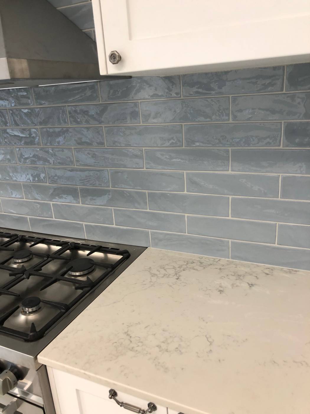 Pasha Steel Blue Gloss 75x300mm tiles installed as a kitchen splashback behind a stainless steel gas cooktop with white marble countertop and white cabinetry.