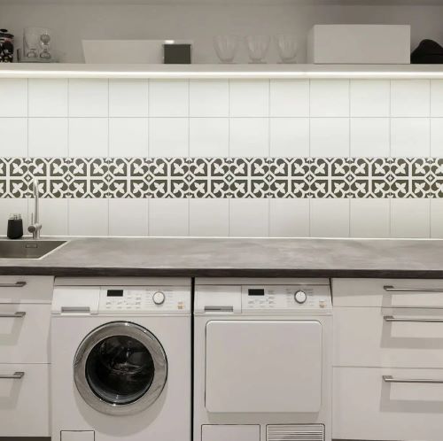Picasso Bloom Black 200x200mm decorative border tiles installed as a horizontal band between white wall tiles and grey countertop in a modern laundry room with washing machines.