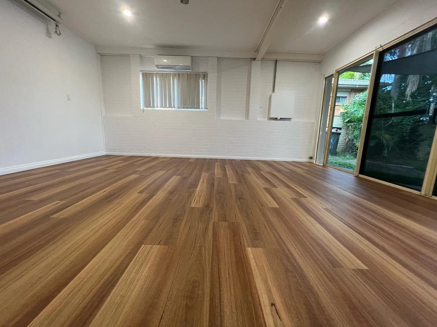 Queensland Spotted Gum Elegant SPC Hybrid 6.5mm Flooring 5+1.5mm installed in an empty living room with white painted brick walls, split-system air conditioning, and sliding glass doors opening to a garden.