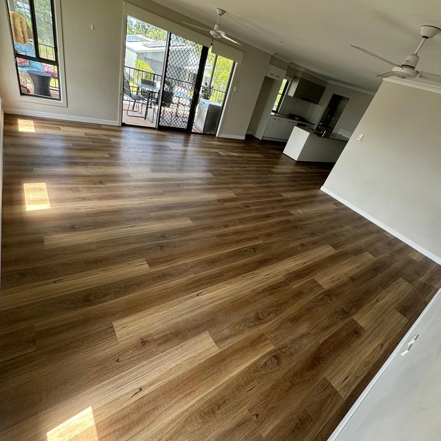 Queensland Spotted Gum Elegant SPC Hybrid 6.5mm Flooring 5+1.5mm installed across a sunlit open-plan living area with ceiling fans and sliding door access to an outdoor deck.