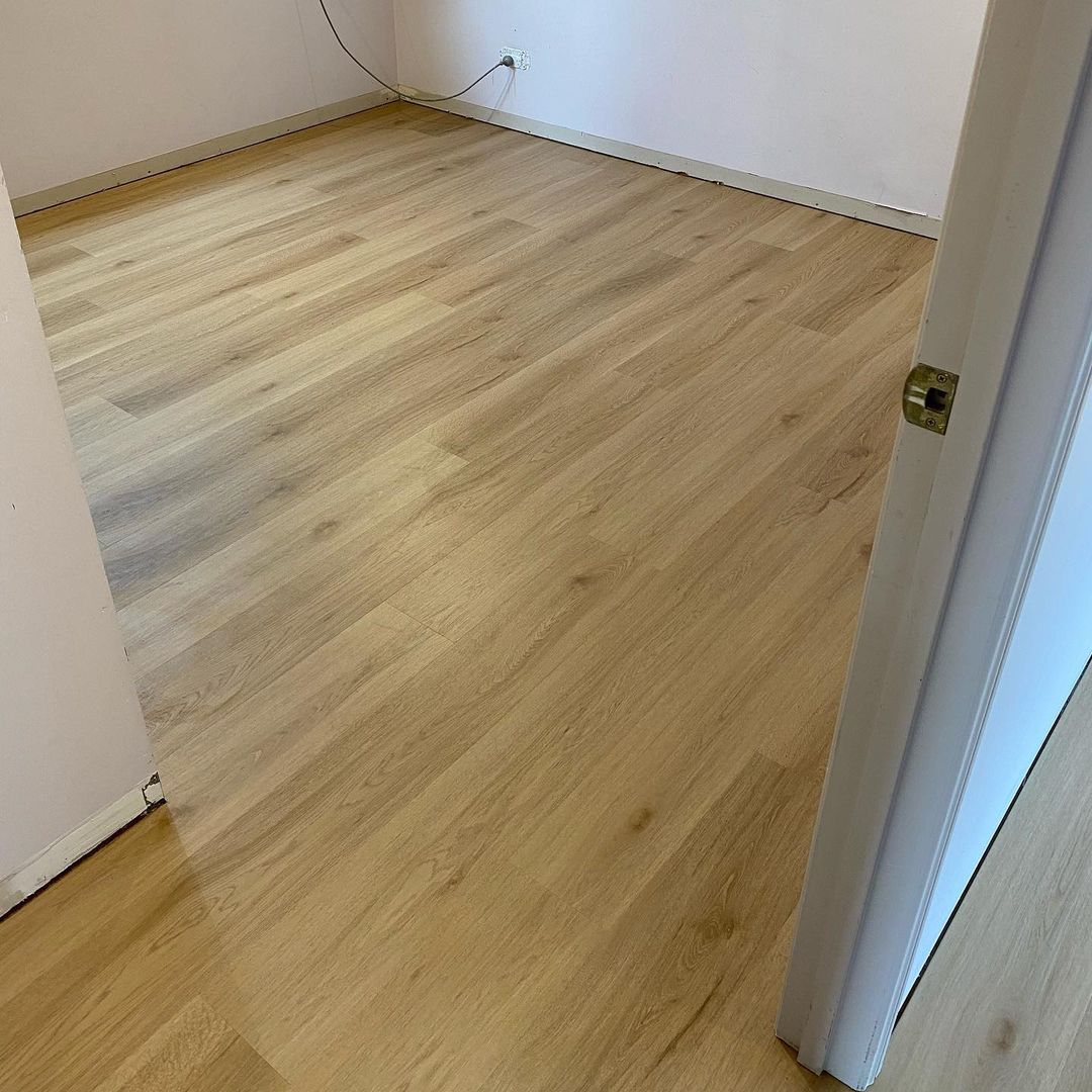 Raw Neutral Elegant SPC Hybrid 6.5mm Flooring 5+1.5mm installed in an empty bedroom, showcasing warm light oak wood-look planks with white skirting boards and a doorway entry.