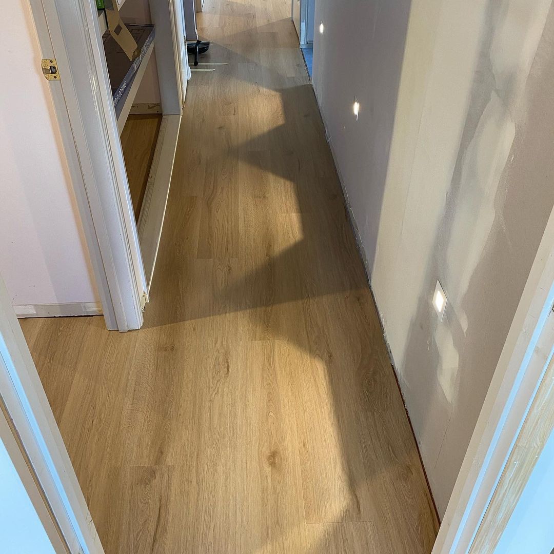 Raw Neutral Elegant SPC Hybrid 6.5mm Flooring 5+1.5mm installed in a narrow hallway with warm oak-look planks, white door frames, and recessed wall lighting.