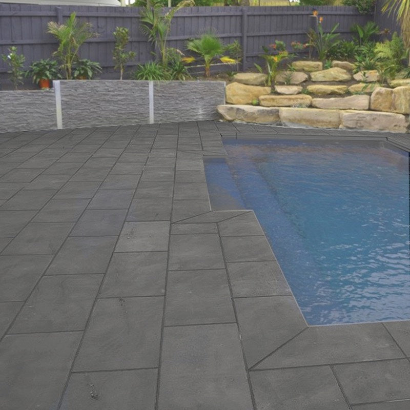 River Stone Dark Grey 300x600mm tiles installed as a pool surround and patio, featuring dark charcoal stone-look pavers edging a blue swimming pool with sandstone garden wall beyond.