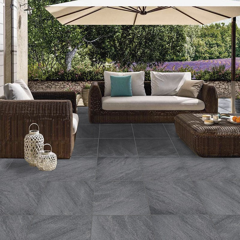 River Stone Dark Grey External Rectified Paver 600x600x20mm installed on an outdoor patio featuring wicker lounge furniture, patio umbrella, and lush garden backdrop.