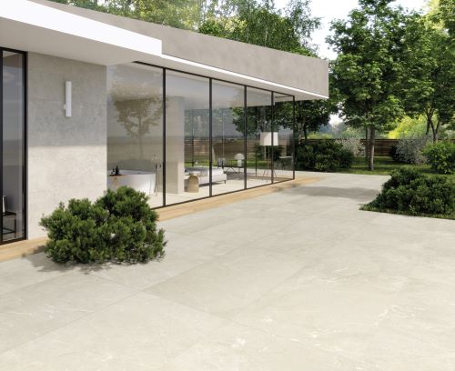 Riviera Ivory 600x600mm porcelain tiles laid on a spacious outdoor patio terrace beside a modern glass-fronted home with lush garden surrounds.