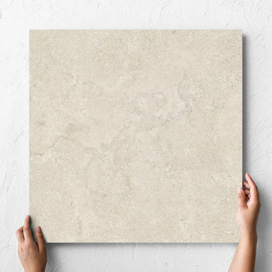 Hand holding a single Roman Travertine Beige 600x600mm tile against a white rendered wall, showing the beige matt surface