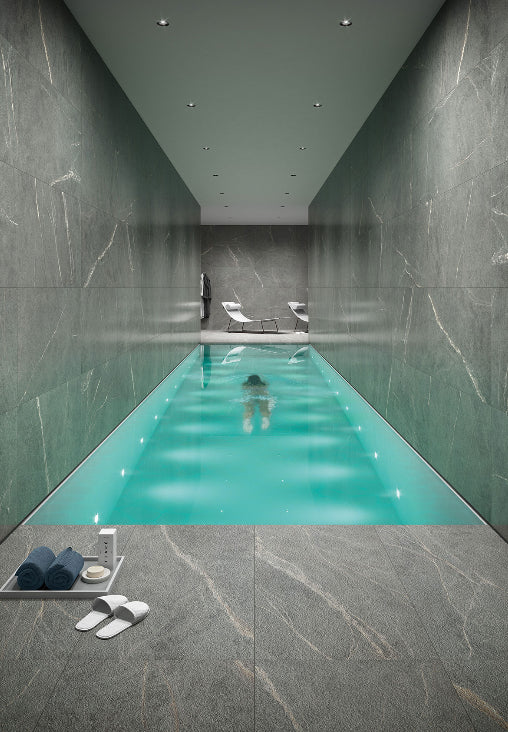 Soap Stone Grey 600x600mm tiles clad the walls and floor of a luxury indoor lap pool with recessed lighting and lounge chairs.