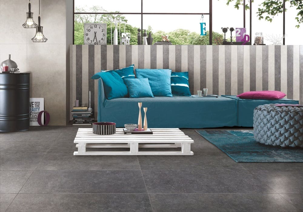 Stella Charcoal Lappato 600x600mm tiles installed as a living room floor, paired with a teal sofa, white pallet coffee table, and industrial pendant lights.