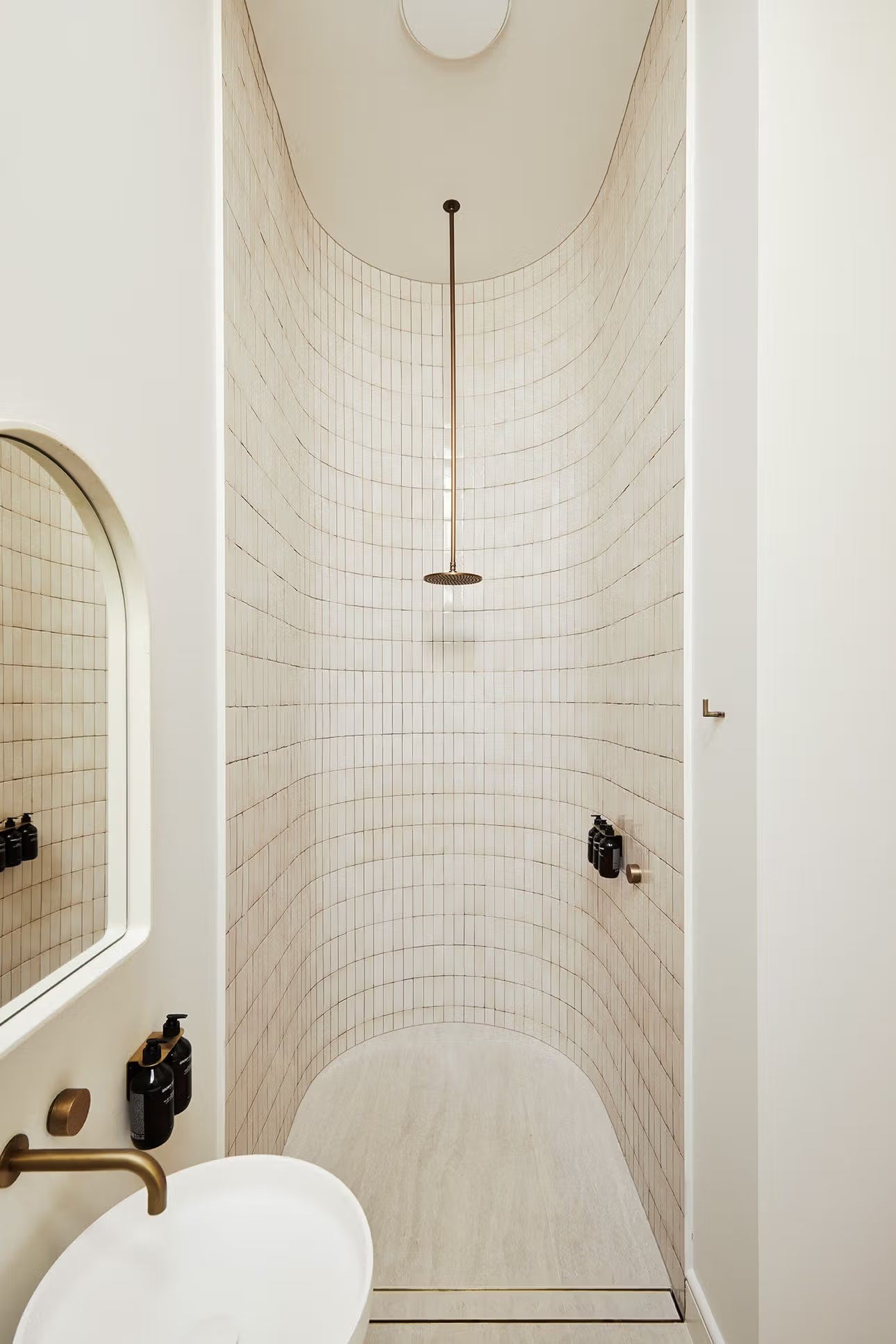 Tallow Vein Cut Travertine Look Ivory 300x600mm tiles lining a curved shower recess with arched ceiling detail, brass pendant rain head, and wall-mounted dispensers.