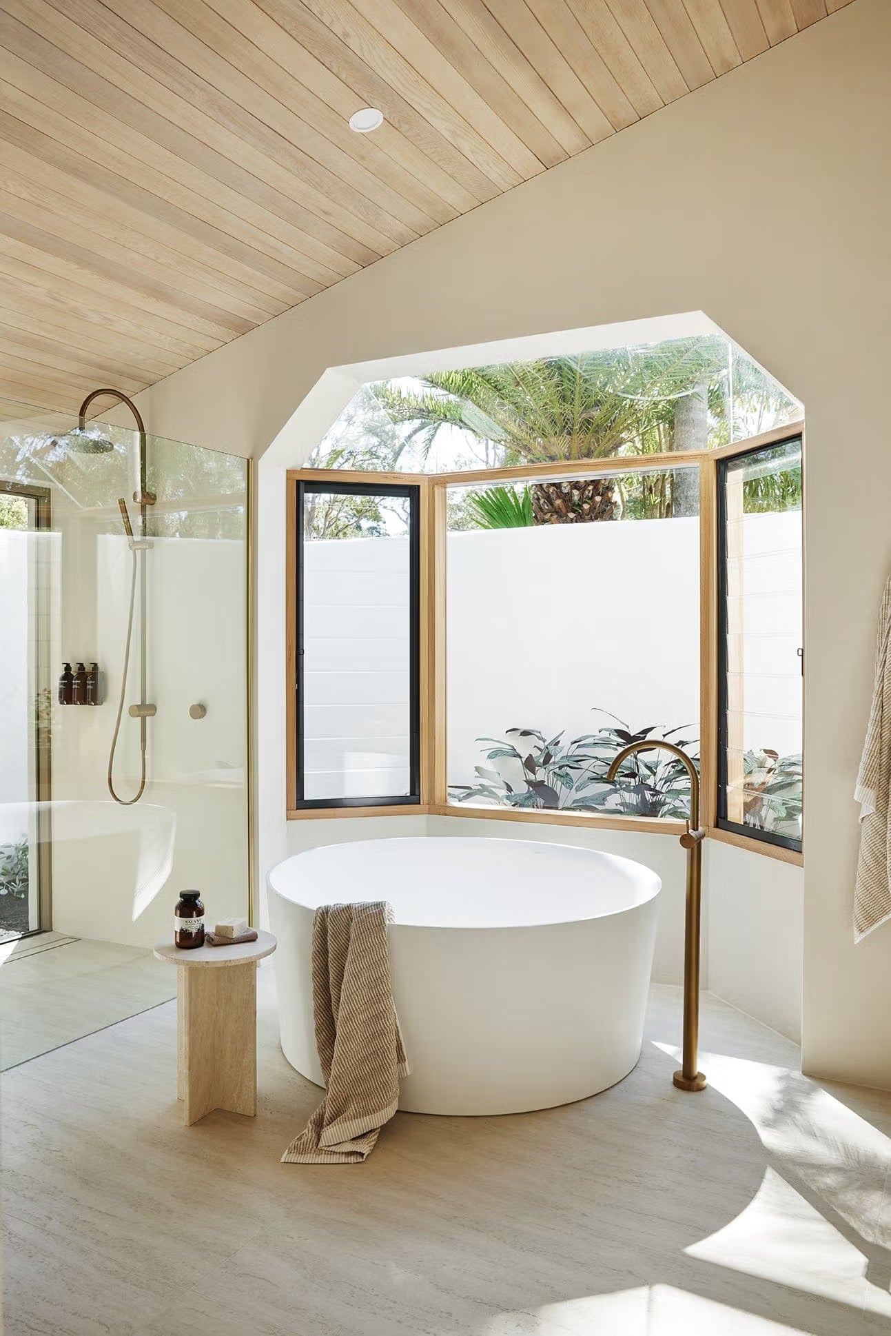 Tallow Vein Cut Travertine Look Ivory 300x600mm floor tiles in a serene spa bathroom featuring a round freestanding bath, brass fixtures, and garden window views.