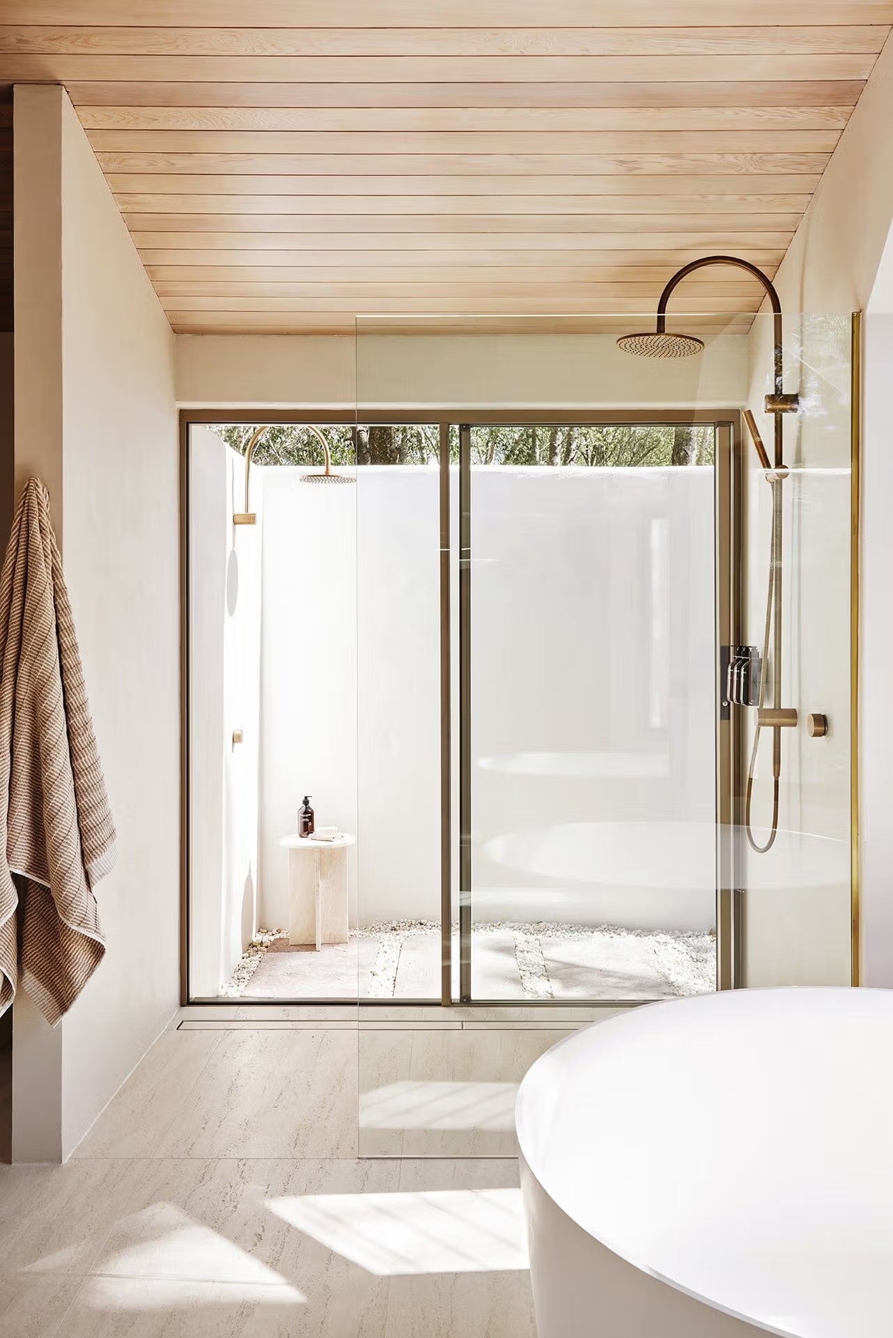 Tallow Vein Cut Travertine Look Ivory 300x600mm tiles laid on a serene bathroom floor leading to a glass-screened indoor-outdoor shower with brass fittings and timber ceiling.
