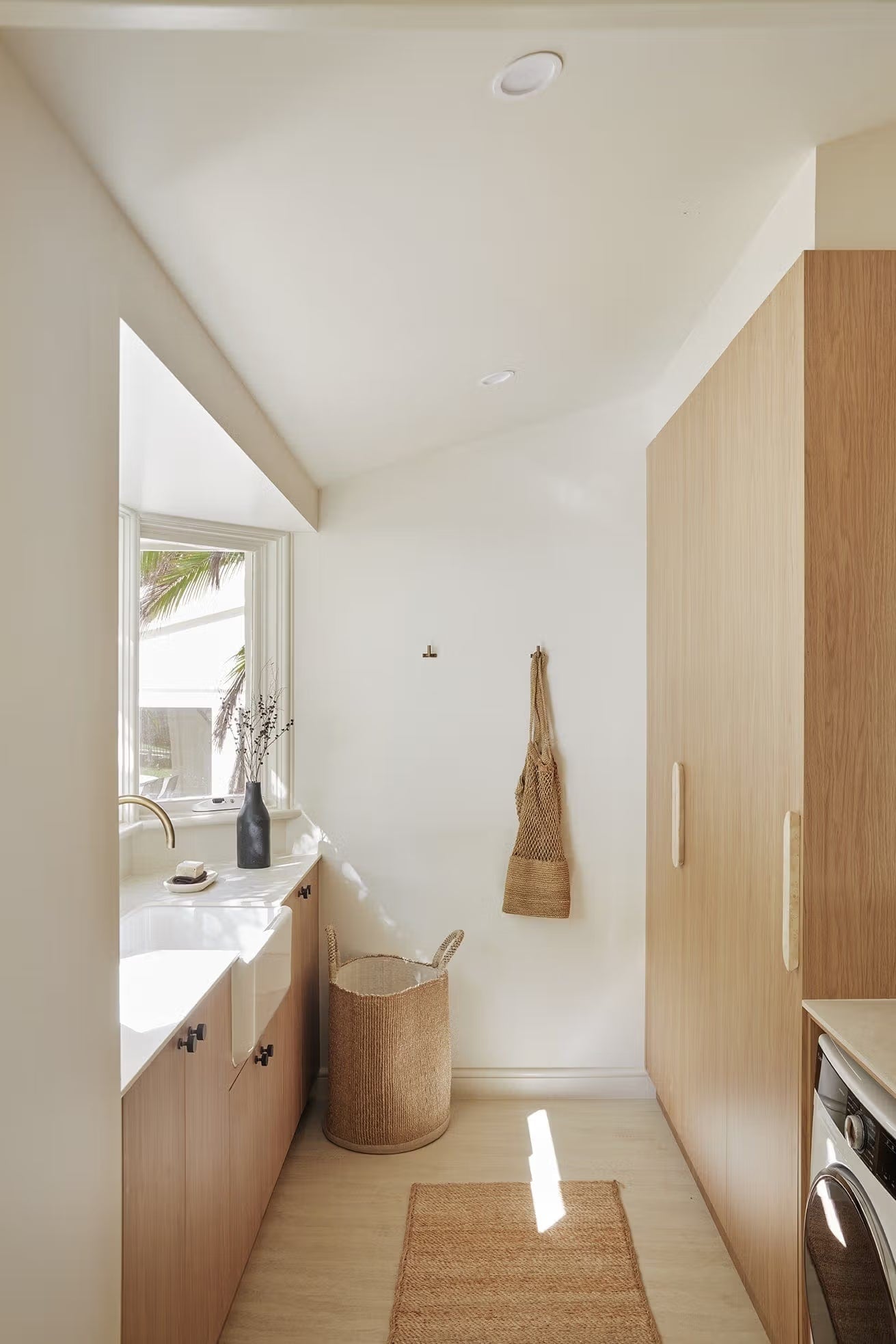 Tallow Vein Cut Travertine Look Ivory 300x600mm tiles on a bright laundry floor paired with warm timber cabinetry, a farmhouse sink, and woven natural-fibre accessories.