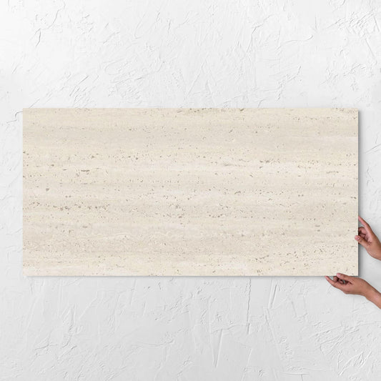 Hands holding a Tallow Vein Cut Travertine Look Ivory 600x1200mm tile against a white rendered wall, showing the beige matt surface