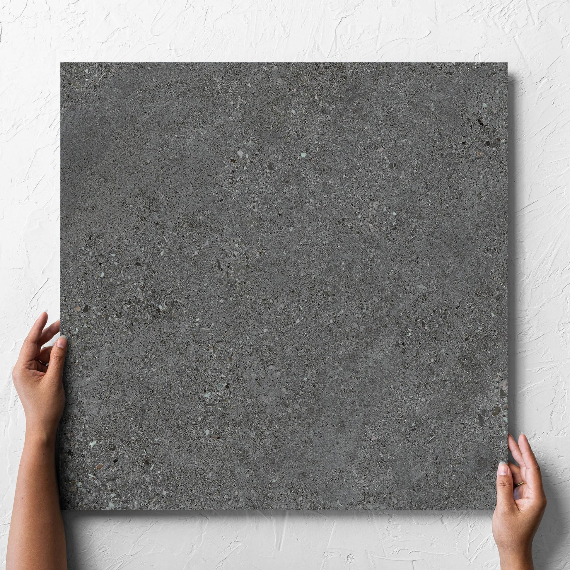 Hand holding a single Terramix Charcoal Grip 600x600mm tile against a white rendered wall, showing the matt surface