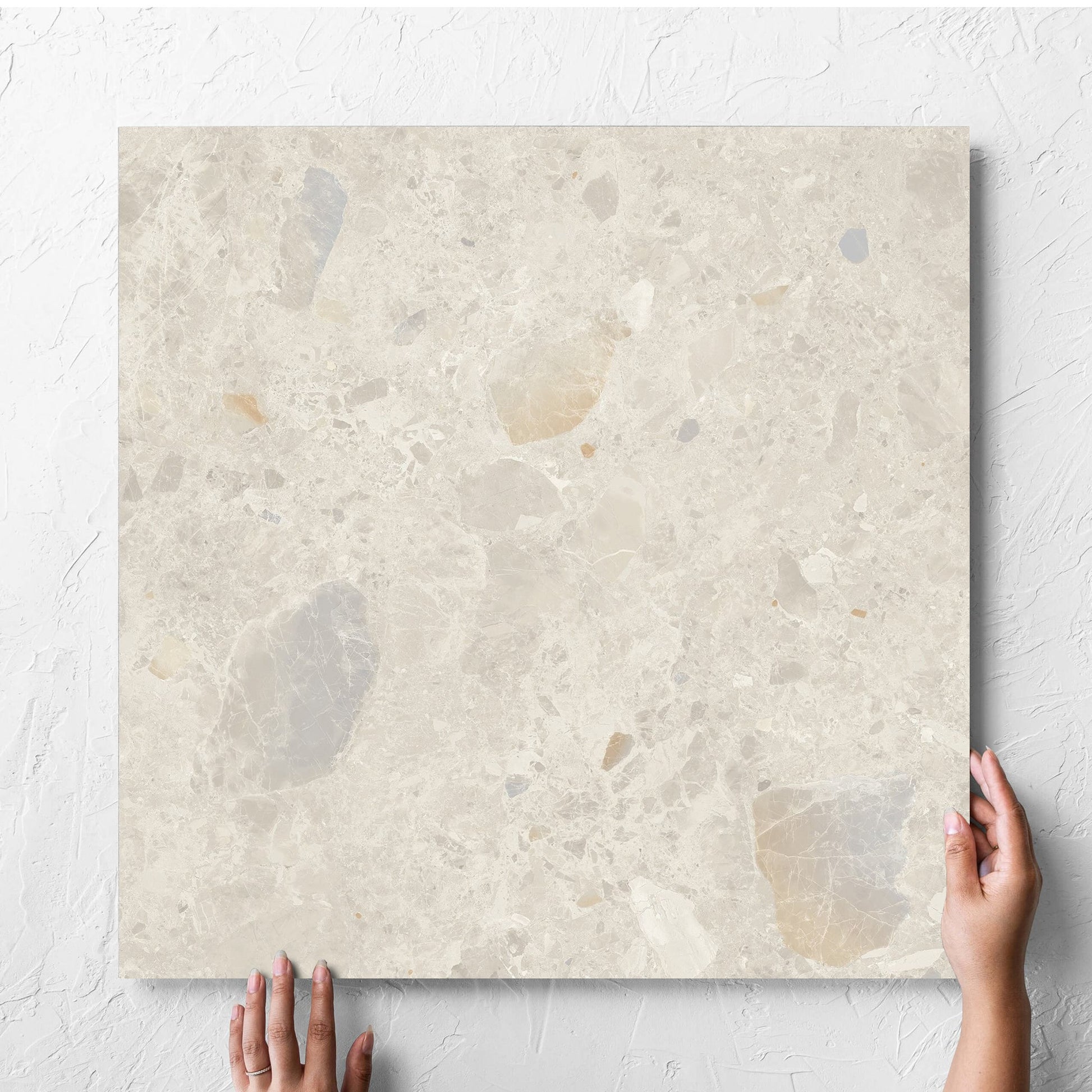 Hand holding a single Terrazzo Rock Beige Matt 600x600mm tile against a white rendered wall, showing the tile surface