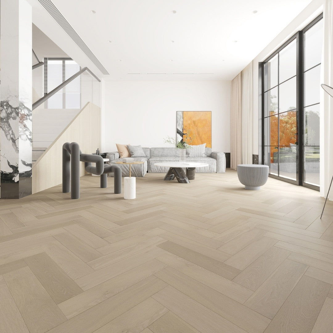 Tinge Clay iDesign Herringbone Flooring 15/3mm installed in a bright open-plan living room with a grey sofa, marble coffee table, and floor-to-ceiling windows.